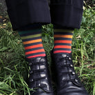 A close-up of a person's legs standing in grass, wearing black cuffed trousers, black lace-up shoes, and highly colorful striped socks with navy, green, orange, red, and yellow bands.