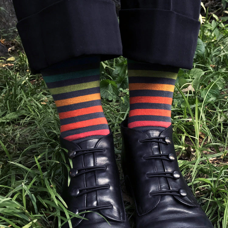 A close-up of a person's legs standing in grass, wearing black cuffed trousers, black lace-up shoes, and highly colorful striped socks with navy, green, orange, red, and yellow bands.