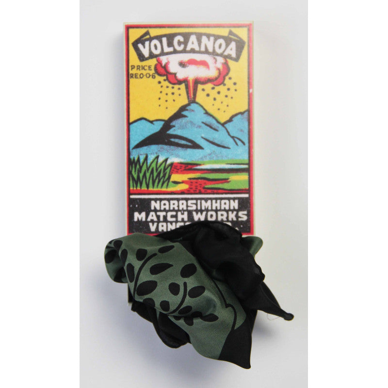 A folded black and olive green patterned scarf or bandana is placed in front of a colorful matchbox-style card that reads 'VOLCANOA' and 'NARBSIMHAN MATCH WORKS'.